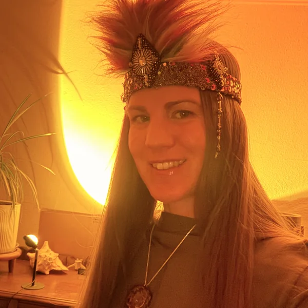 Cassandra Beaumont wearing a beaded headdress with a feather plume, lit by warm lamplight.