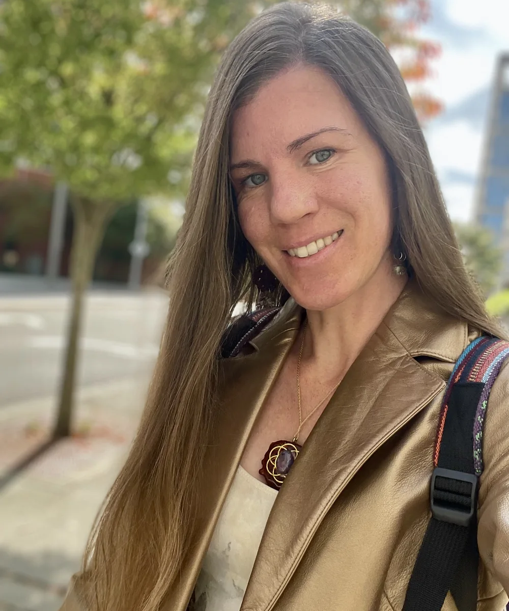 Cassandra Beaumont, smiling outdoors in a gold leather jacket, wearing a sacred-geometry pendant.